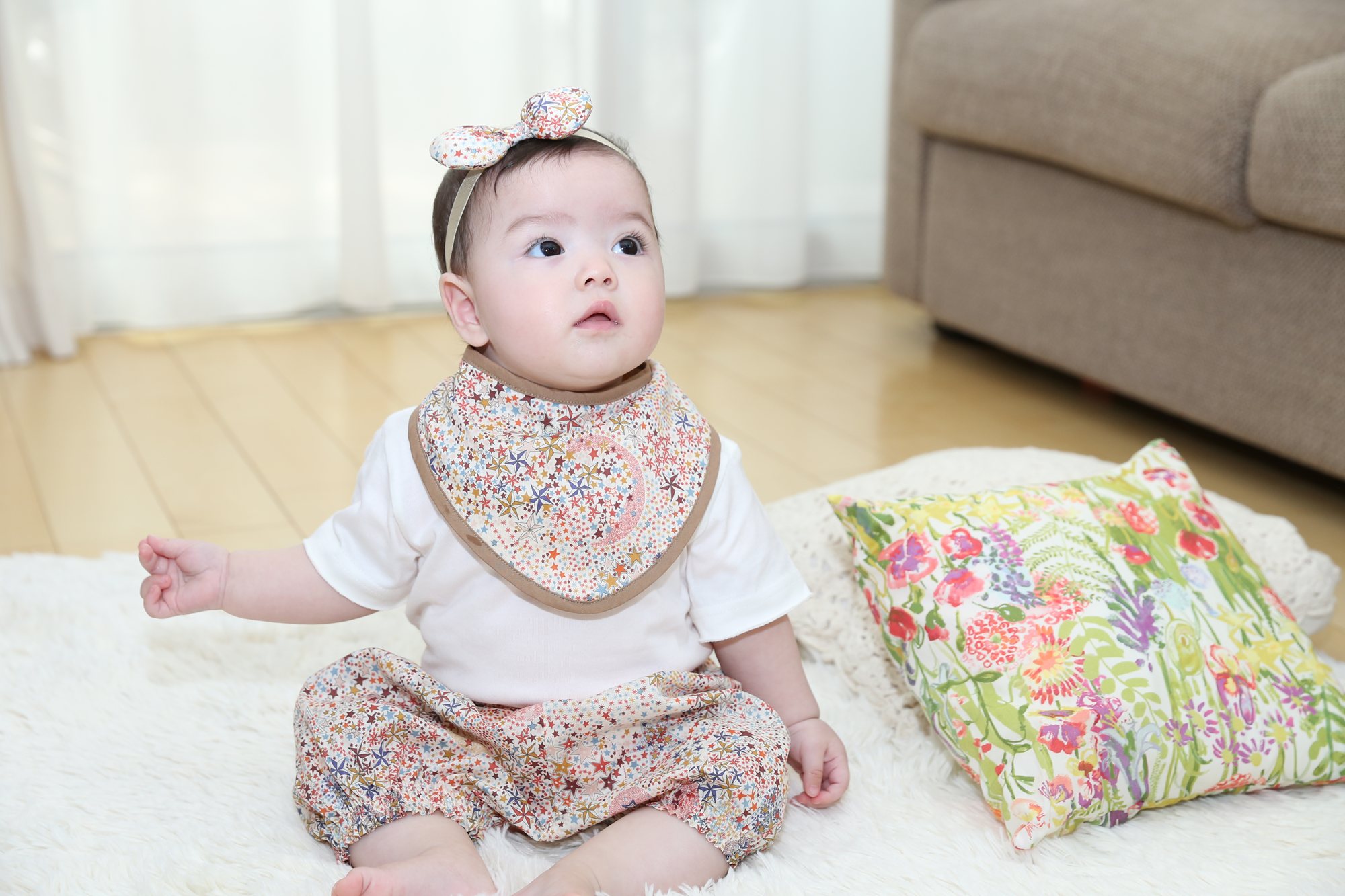 リバティ柄のスタイとヘアバンド、おそろいのパンツを身に着け、白いラグの上でお座りする赤ちゃん