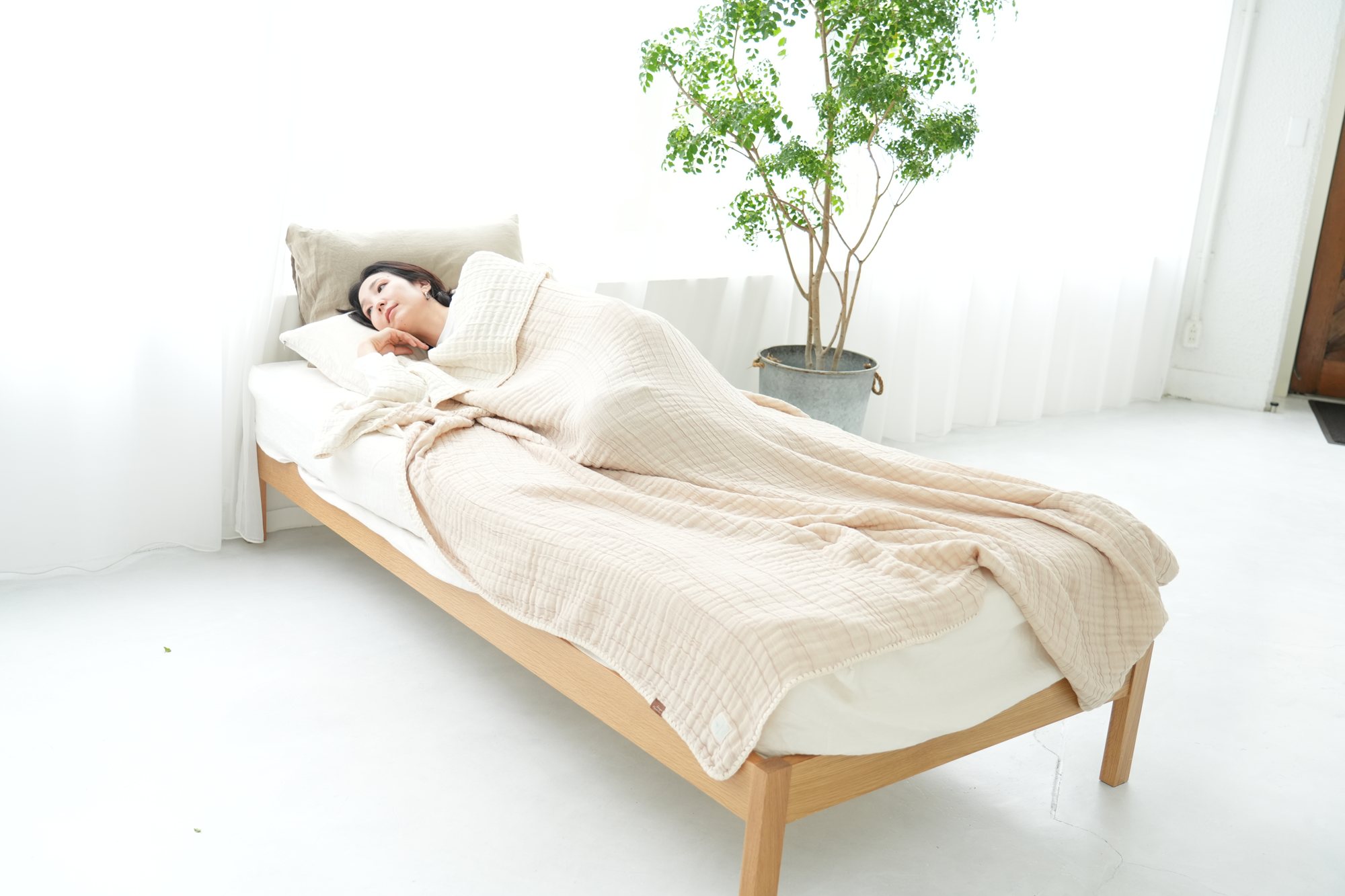 観葉植物のある白く明るい寝室で、ベージュの四重ガーゼケットを掛けてベッドに眠る女性