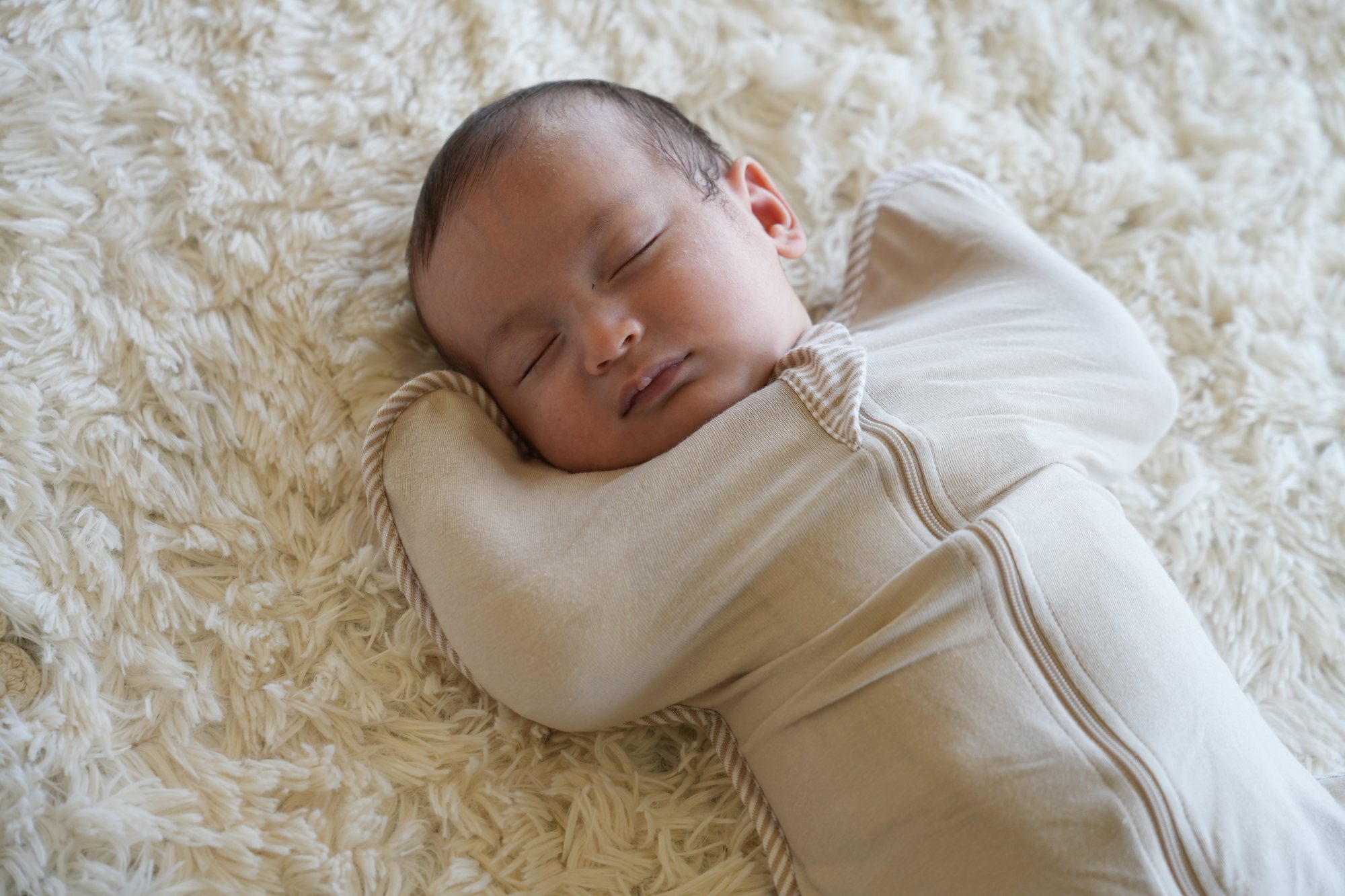 ベージュのスワドルに包まれて、両手を顔のそばに上げたまま眠る赤ちゃんの顔まわりのアップ