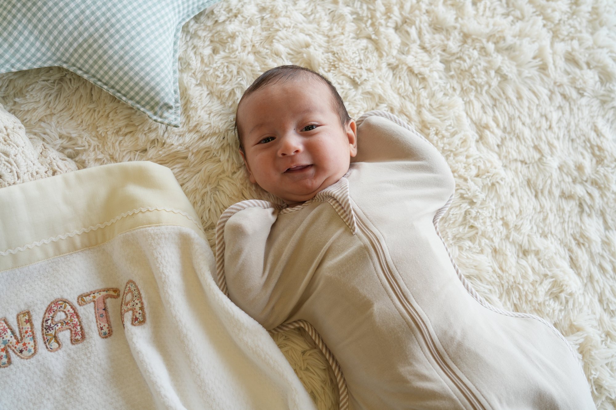 ベージュのスワドルを着てボアラグに寝た赤ちゃんがにっこり。手前には名前入りのタオルケットがのぞいている