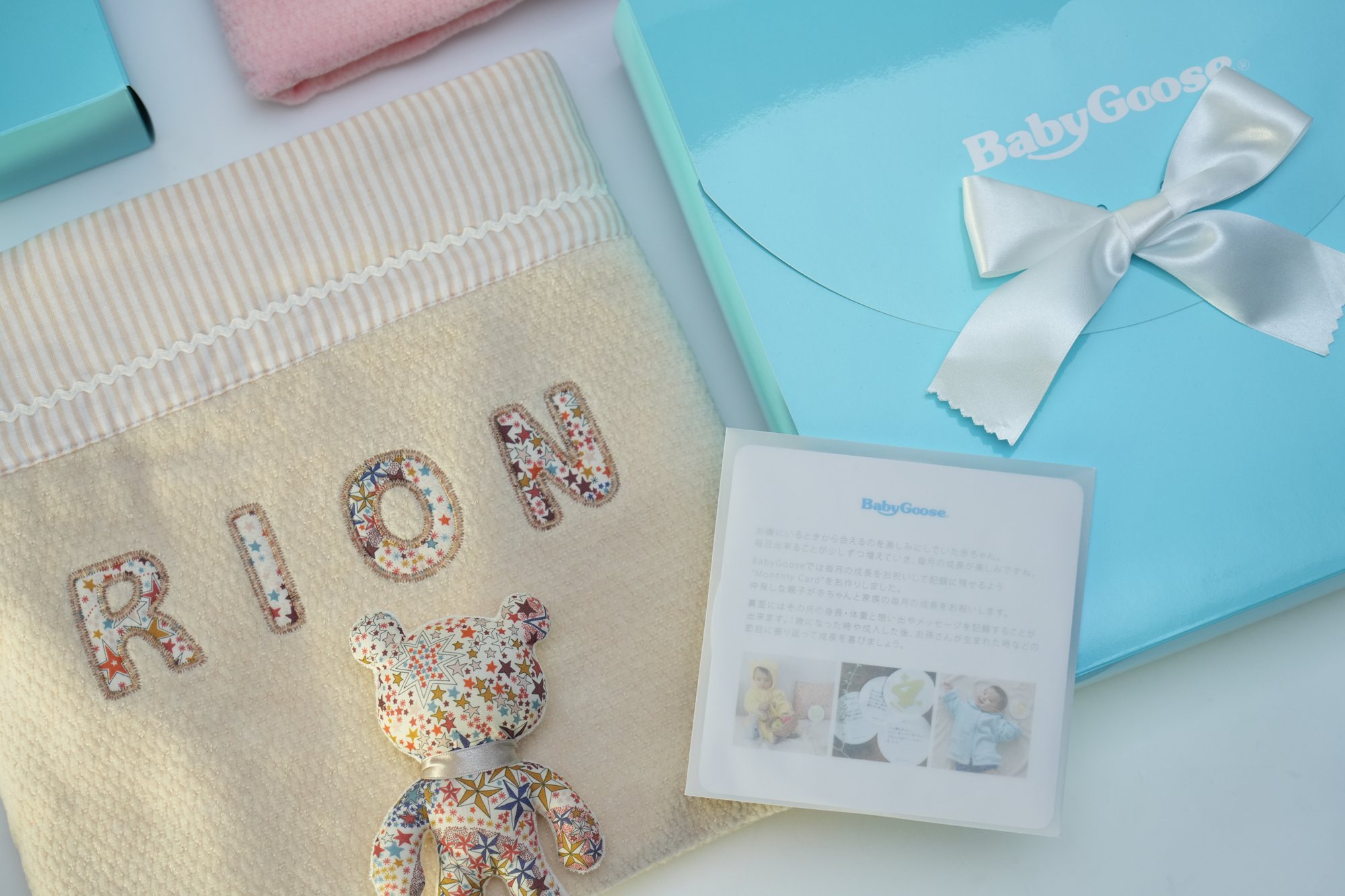 RIONの名入れタオルケットに小花柄のくまのアップリケ、BabyGooseのカードと水色のギフトボックスを添えた寄り