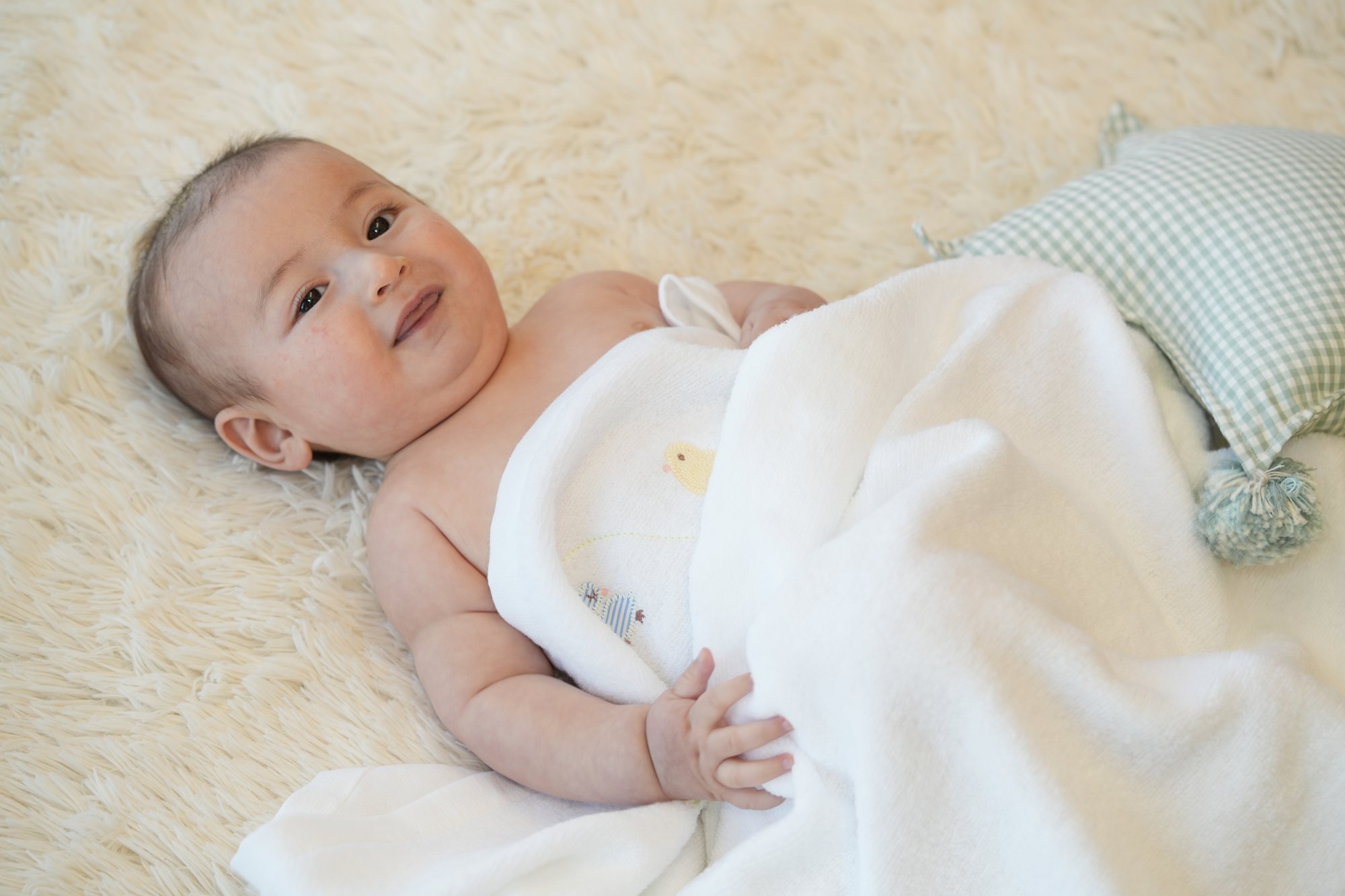 ひよこのワンポイントが付いた白いケットを掛けて、ファーラグの上でにっこり笑う赤ちゃん
