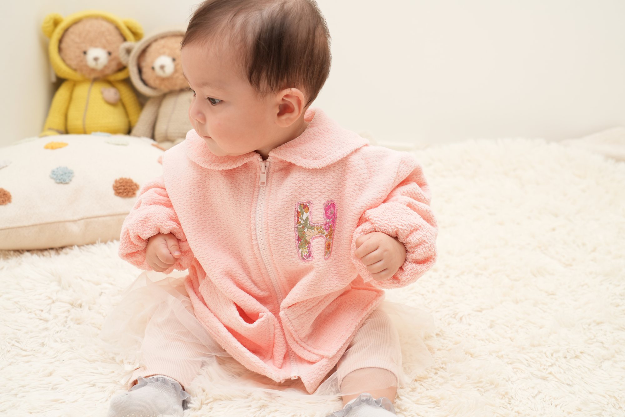 胸元に花柄のHのアップリケが入ったピンクのジャンパーの女の子が、白いラグに座って横を向いている