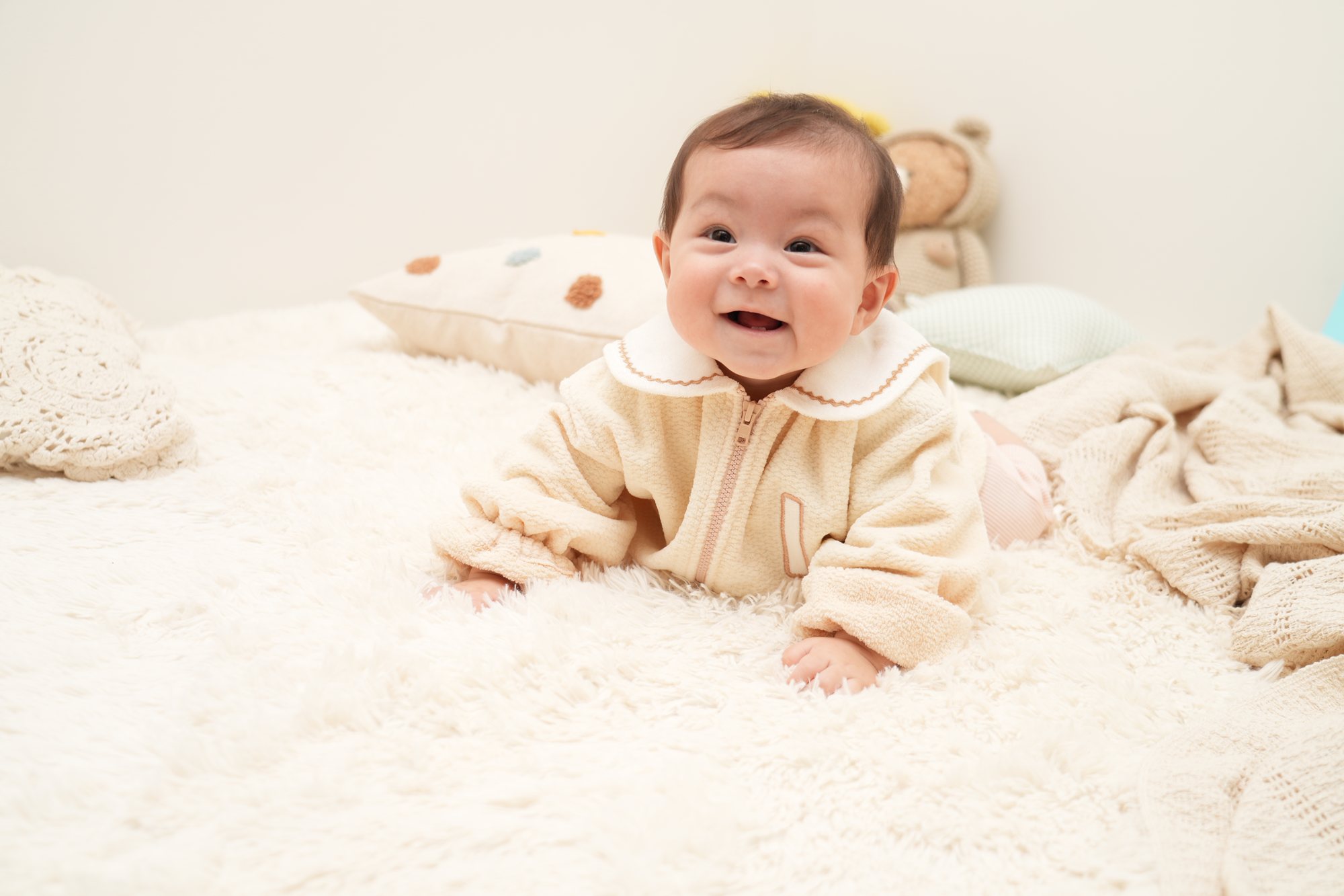 セーラー衿のアイボリーのジャンパーを着た赤ちゃんが、白いふわふわのラグにうつ伏せで顔を上げて笑っている
