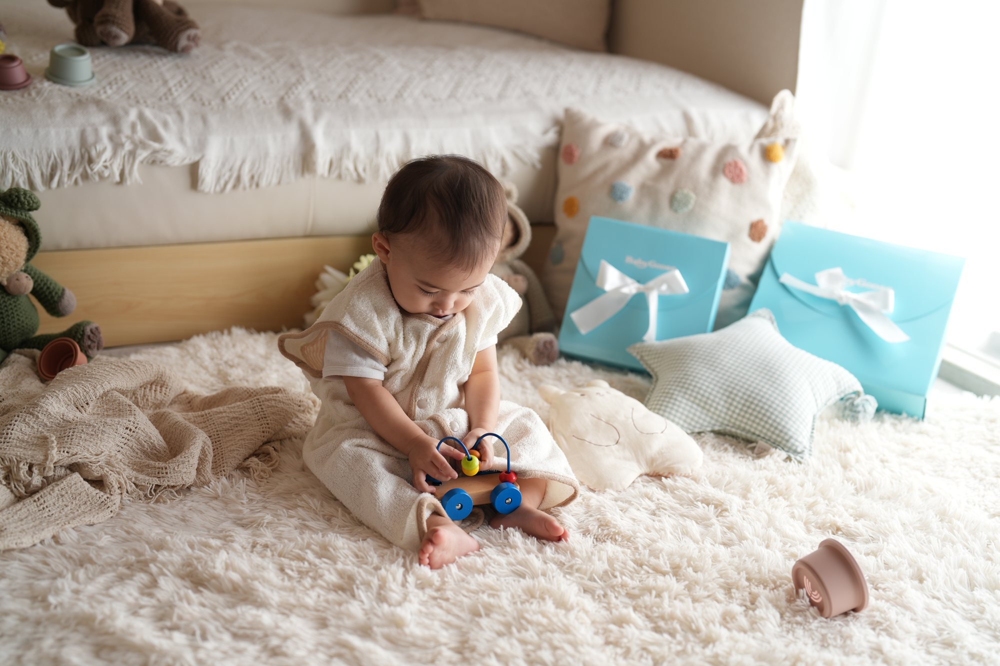 生成りのガーゼの服で白いラグに座り、カラフルな木のおもちゃで遊ぶ赤ちゃん。奥に水色のギフト箱