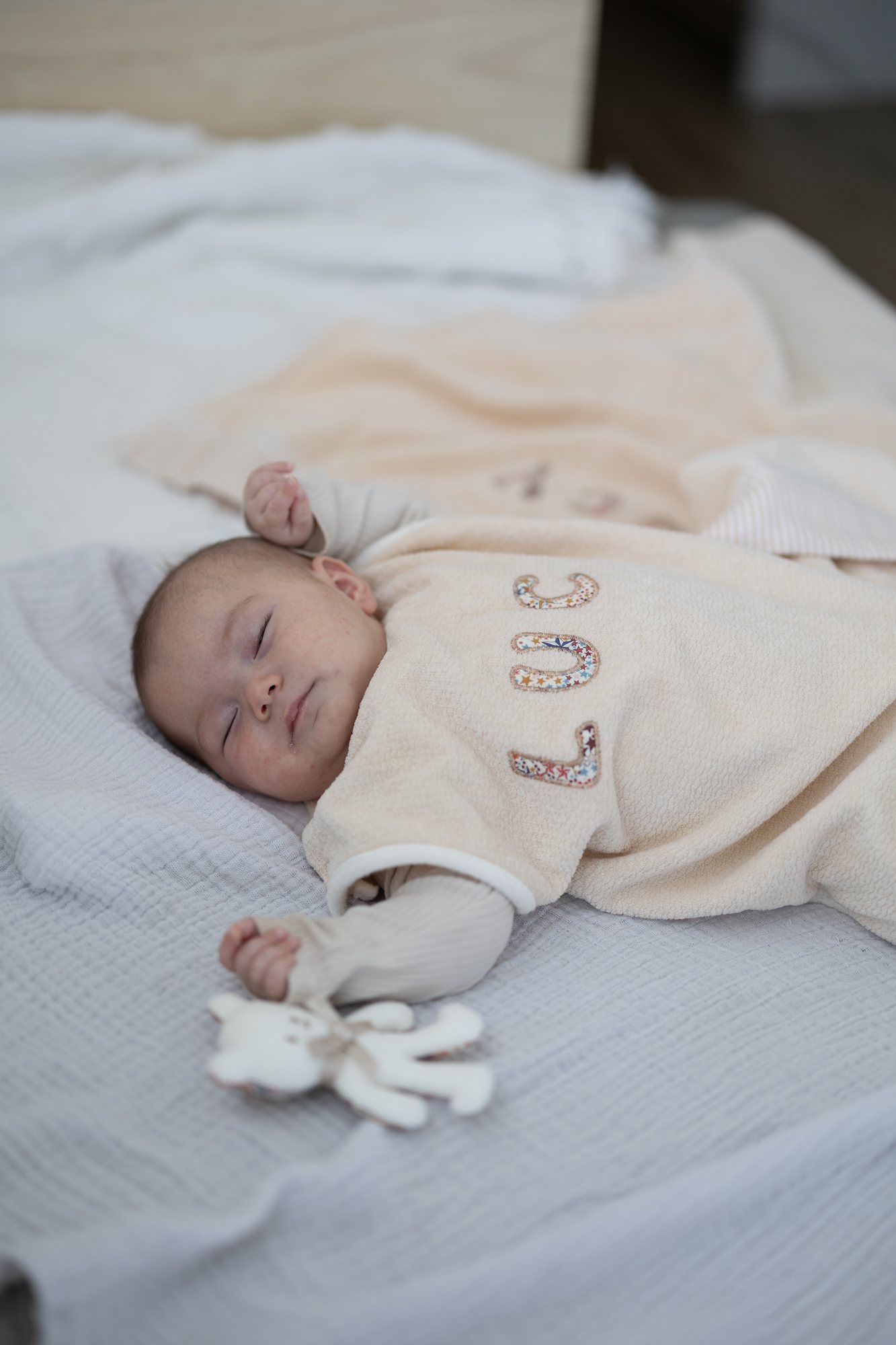 胸に小花柄の名前が入った生成りのスリーパーを着て、ガーゼケットの上で眠る赤ちゃん。奥にも名入れのケットが見える