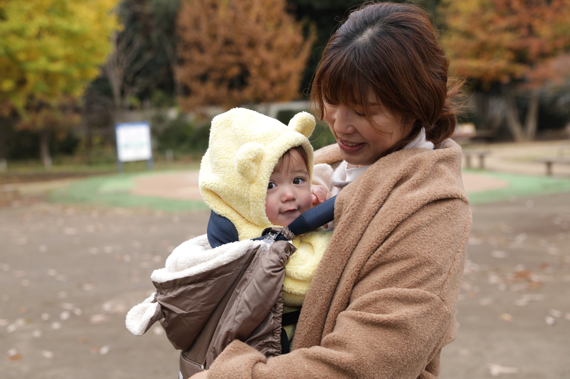 防寒ケープ付きの抱っこ紐で、くまフードの赤ちゃんに頬を寄せてほほえむ大人、紅葉の公園で