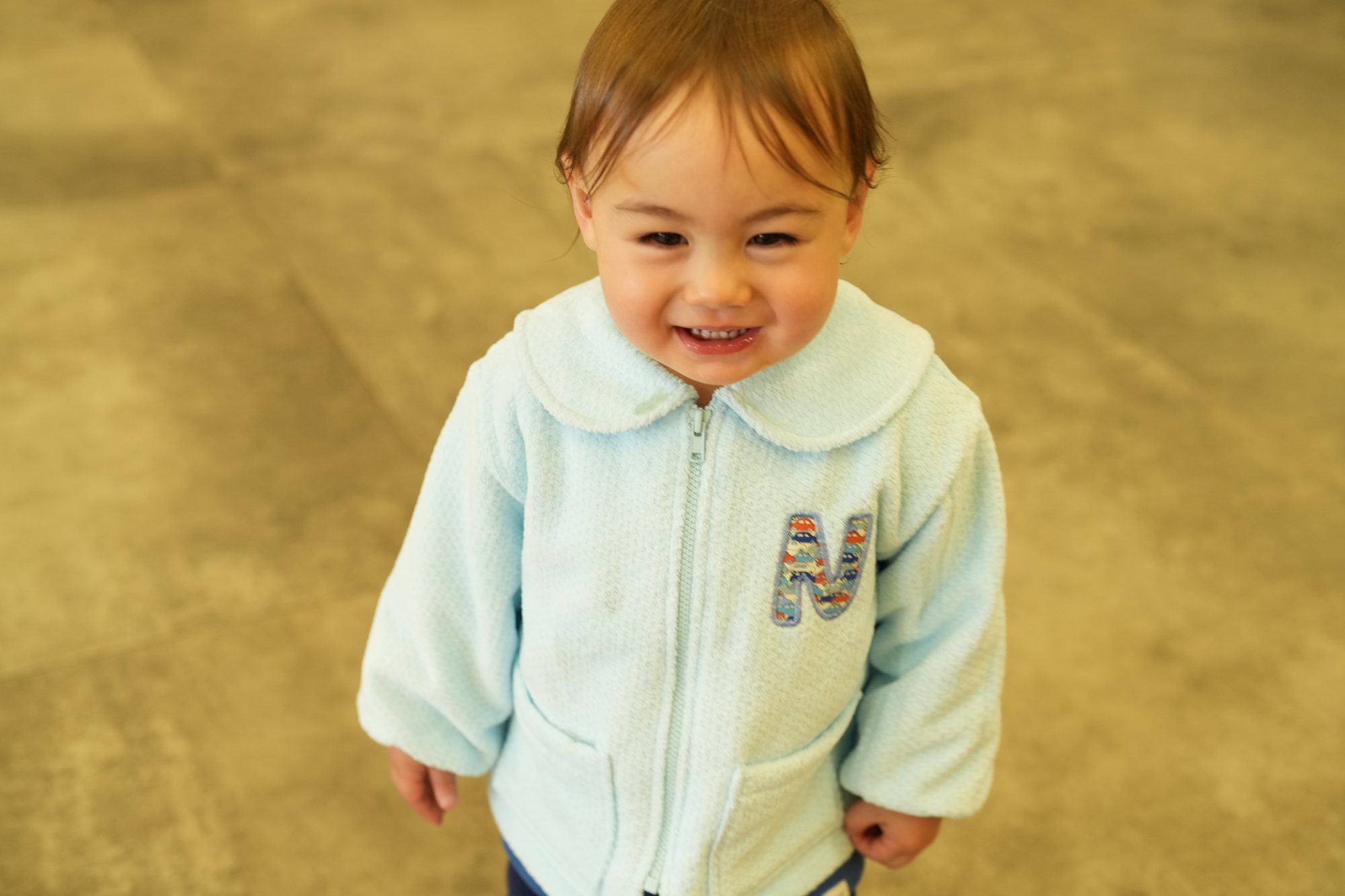 胸にイニシャルのアップリケが入った水色のジャンパーを着た子が、顔を上げて目を細めてにっこりしている