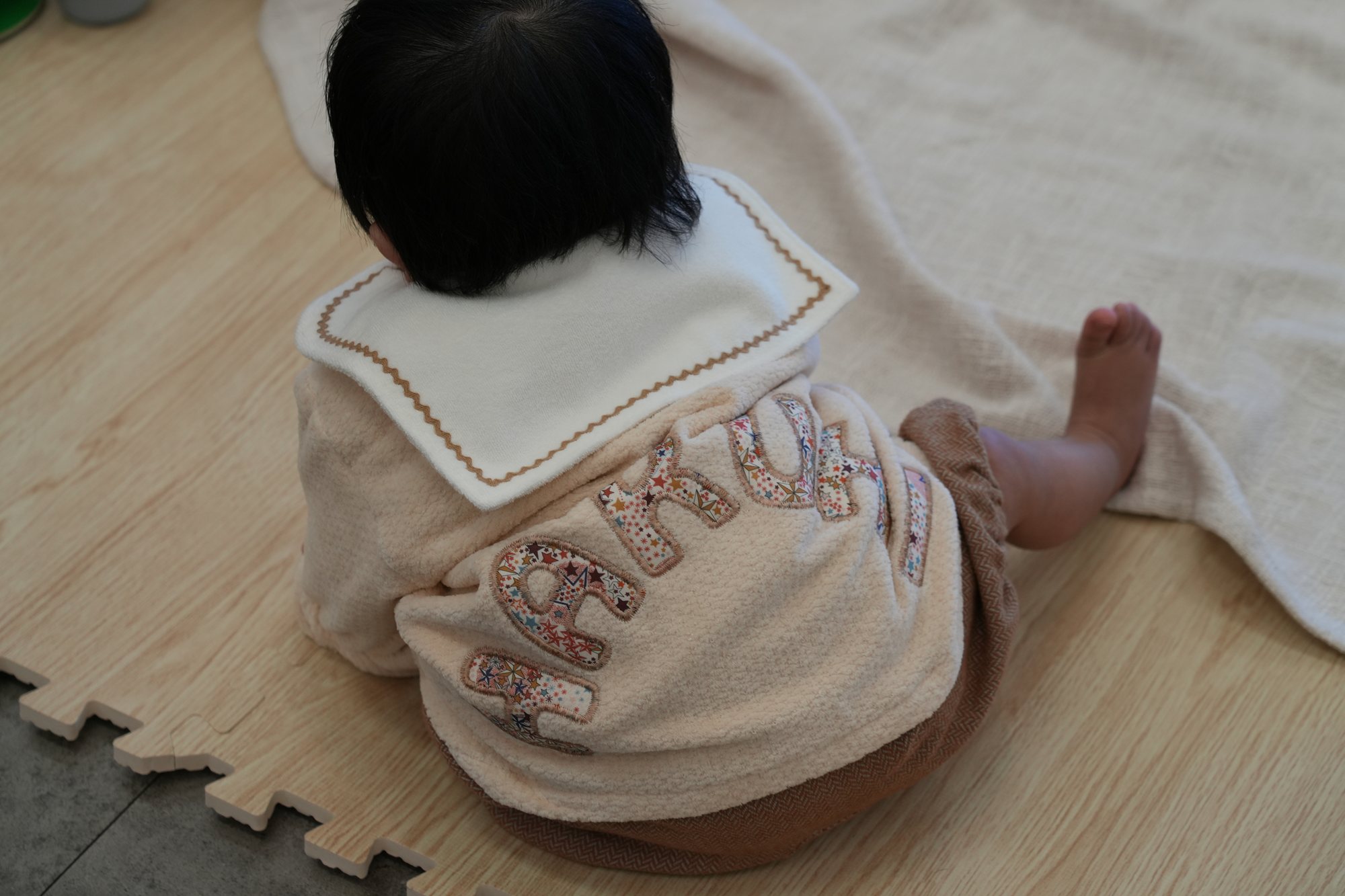セーラー襟のベージュのジャンパーを着て座る赤ちゃんの後ろ姿、背中にリバティ柄の名前アップリケ