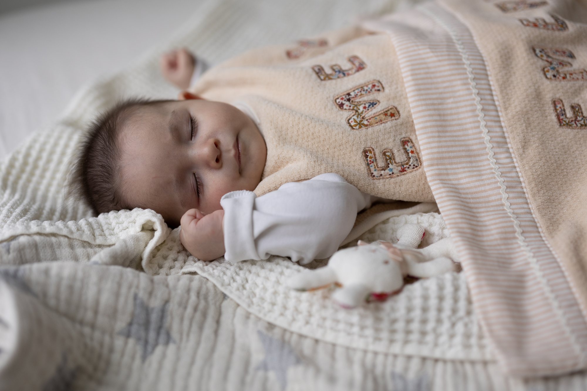 ワッフルケットの上で横向きにすやすや眠る赤ちゃんと、名前アップリケ入りのベージュのケット、うさぎのぬいぐるみ