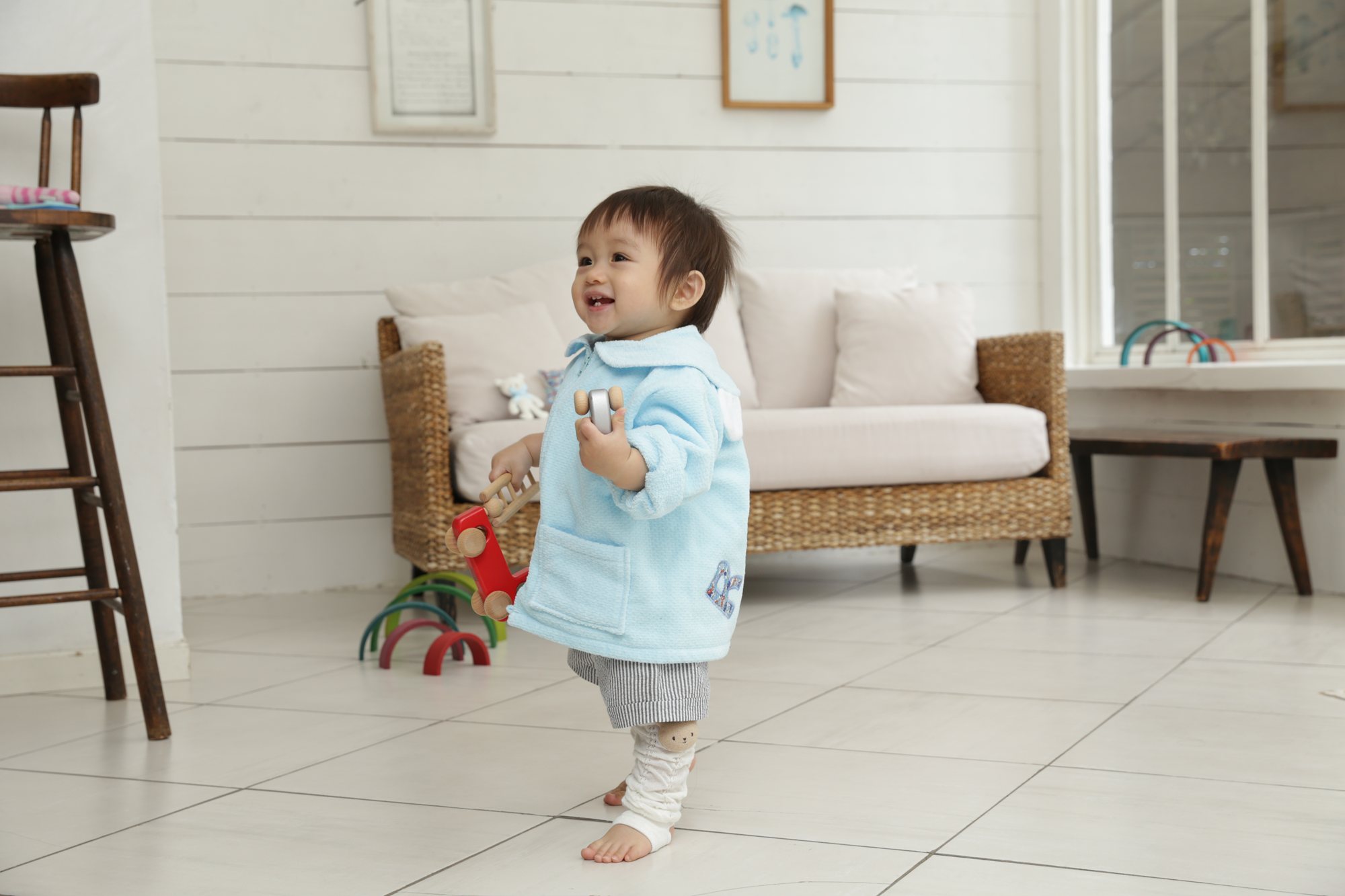 水色のジャンパーを着た1歳くらいの男の子が、白い部屋でおもちゃを手に、笑いながらこちらへ歩いてくる