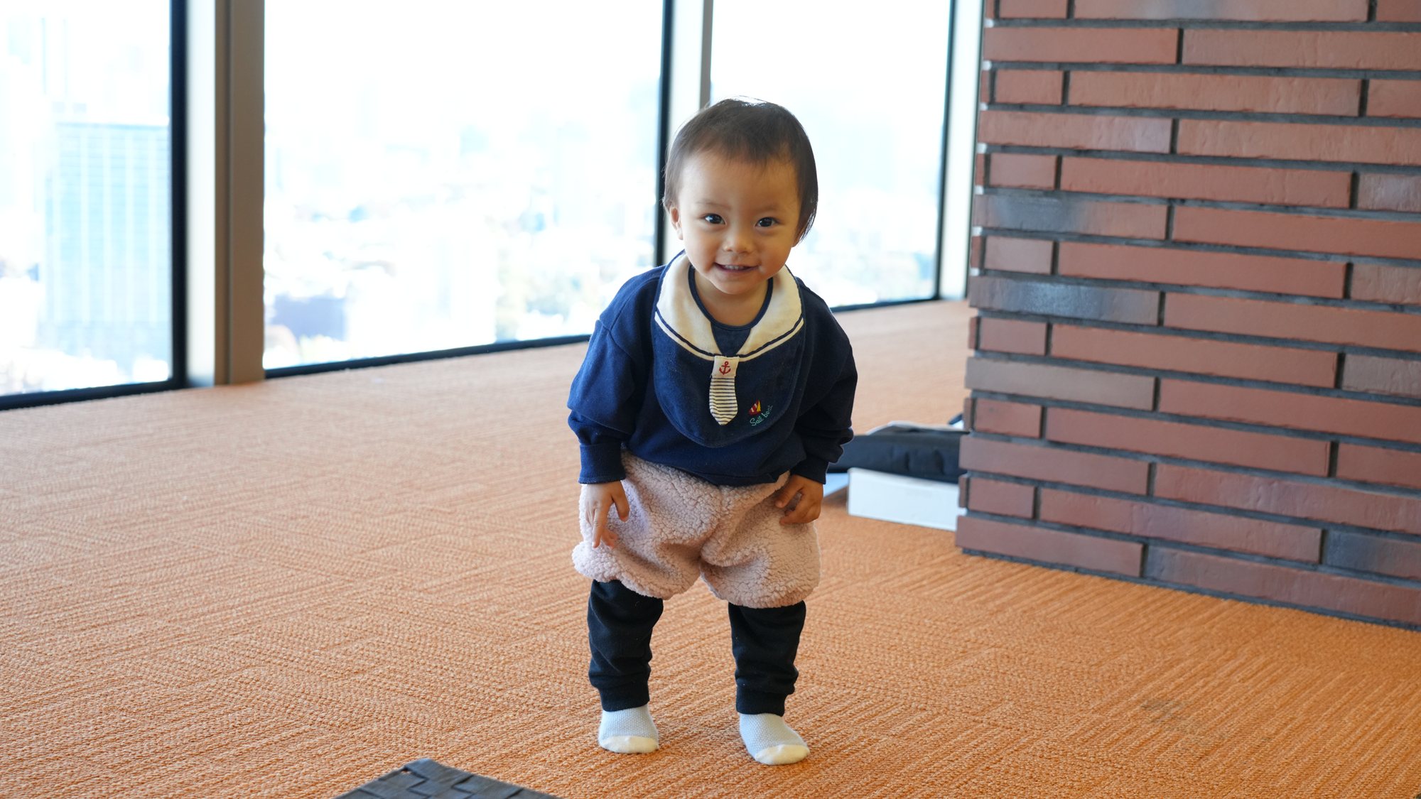 紺のセーラー襟のジャンパーを着て、高層階の窓辺のカーペットに立ちにっこり笑う子ども