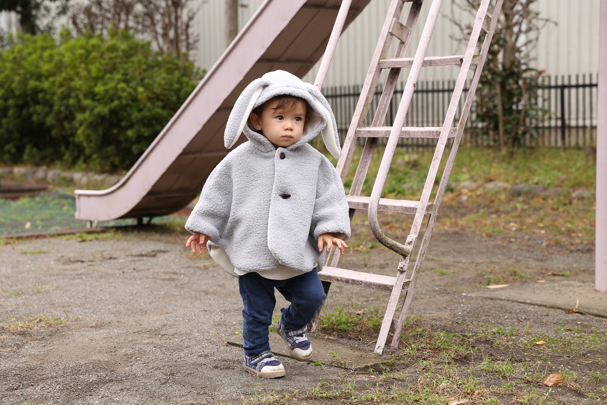 うさぎ耳フードのグレーのボアポンチョを着て、公園のすべり台のそばをとことこ歩く男の子。来年の冬にはこんなお外遊びが楽しみに